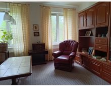 Ein Wohnzimmer mit einem großen Fenster mit gelb karierten Vorhängen, einem braunen Ledersessel, einem hölzernen Wohnzimmerschrank, einem rechteckigen Holztisch und einem Teppichboden. Auf der linken Seite steht eine Pflanze in einem Korb, und an der Wand hängt ein kleines Radio auf einem Holzregal.