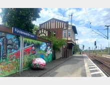 Bahnhof Fürstenwalde (Spree) mit einem gemauerten Stellwerksgebäude im Fachwerkstil rechts und einer mit buntem Graffiti bemalten Wand links, die eine Naturszene und einen roten Zug zeigt. Im Hintergrund sind Gleise, Oberleitungen und Signalanlagen sichtbar.
