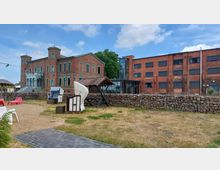 Historisches Backsteingebäude mit Türmen und grünen Fensterrahmen, daneben ein modernes Backsteingebäude mit großen Fenstern und Glasfassade; im Vordergrund ein gepflasterter Weg, ein Sandbereich mit Strandkörben, Holztischen und Trockenrasen, umgeben von einer niedrigen Steinmauer.