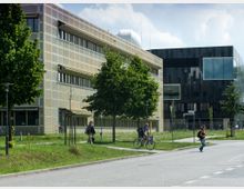 Moderne Gebäude in einer urbanen Umgebung mit großen Fenstern und einer Fassade aus Metall und Glas. Im Vordergrund eine Grünfläche mit Bäumen, Gehwegen und Passanten sowie einem Radfahrer.