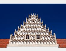 Giebeldetail eines historischen Gebäudes mit Stufengiebel in symmetrischem Fischblasen-Muster und zwei Fenstern, flankiert von roten Ziegeldächern vor klarem, blauem Himmel.