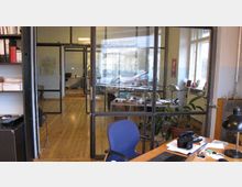 Ein modernes Büro mit Holzboden und Glaswänden, das einen Blick in einen weiteren Arbeitsbereich ermöglicht. Im Vordergrund befinden sich ein Schreibtisch mit Büroartikeln, ein blauer Bürostuhl und Pflanzen in Töpfen, während draußen Autos zu sehen sind.