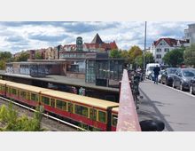 S-Bahn-Station in städtischer Umgebung mit einem roten Zug auf den Gleisen, einem überdachten Bahnsteig mit Graffiti an den Wänden sowie einem Glasaufbau. Im Hintergrund sind ältere Gebäude mit roten Dächern und Bäumen sichtbar, und rechts verläuft eine Straße mit geparkten Autos und Fahrrädern.