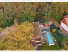 Luftaufnahme eines modernen Holzhauses mit begrüntem Flachdach, einer Terrasse und einem länglichen Außenpool. Das Haus liegt inmitten eines waldreichen Gebiets, mit weiteren Gebäuden und geparkten Autos in der Nähe.