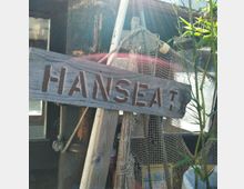 Ein hölzernes Schild mit der Aufschrift „HANSEAT“ ist vor einem rustikalen Hintergrund aus Fischernetzen, Holzpfählen und Bambuszweigen angebracht. Diffuses Sonnenlicht fällt auf die Szene.