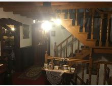 Gemütlicher Innenraum mit einer Holztreppe und Geländern, die zu einer oberen Etage führen. Im Vordergrund steht ein Tisch mit einer alten Nähmaschine und gerahmten Fotos, daneben befinden sich Teppiche und traditionelle Holzmöbel, die den Raum schmücken.