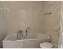 Badezimmer mit beigefarbenen Fliesen, einer rechteckigen Badewanne mit Duscharmatur und Handbrause sowie einer Toilette daneben. Im Hintergrund ist ein Handtuchhalter an der Wand befestigt.