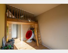 Ein modernes Kinderzimmer mit Hochbett aus Holz und Leiter, darunter eine Tür sowie eine Sitzecke mit einem weißen, kugelförmigen Stuhl mit rotem Innenpolster. Der Raum hat helle Wände, Holzboden und ist mit Spielzeug und Dekorationen ausgestattet.