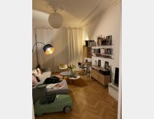 Wohnzimmer mit Parkettboden, einer grauen Couch und dekorativen Kissen, einem Kinderspielzeugauto sowie einem runden Couchtisch. Im Hintergrund stehen Regale mit Büchern, eine kleine Kommode und eine Stehlampe; weißer Vorhang und verzierte Stuckdecke.