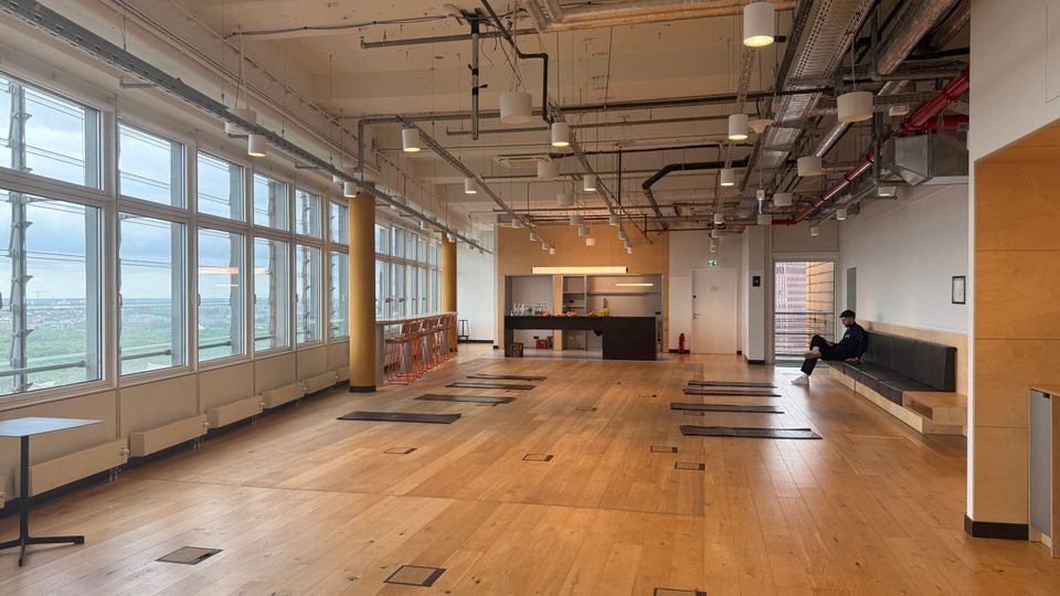 Large open-plan studio or office space with a light wood floor, floor-to-ceiling windows along one side, and an exposed industrial ceiling with visible ducts, pipes, and hanging lights. A small kitchenette/bar area sits at the far end, with high stools by the windows and a long bench against the wall.
