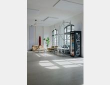 Heller, minimalistischer Loft-Raum mit hohen Decken, großen Rundbogenfenstern und glattem Betonboden. In einer Sitzecke stehen beigefarbene Sofas, ein niedriger Tisch und Pflanzen; daneben ein fahrbares Gerüst und ein Getränkeautomat an der Wand.