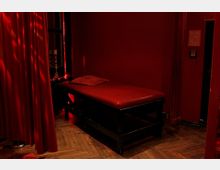 Ein Raum mit roten Wänden, rotem Vorhang und gedämpfter Beleuchtung. Im Vordergrund steht eine gepolsterte rote Liege auf einem dunklen Gestell, der Holzboden ist mit Fischgrätenmuster gestaltet.