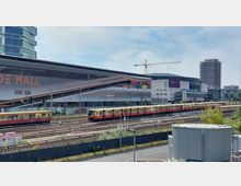 Ein Bahnhof mit gelber S-Bahn und mehreren Gleisen im Vordergrund. Im Hintergrund befinden sich ein modernes Einkaufszentrum mit der Beschriftung „Mall“, weitere Geschäftsgebäude sowie ein Hochhaus in Bau und ein Kran.