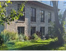 Backsteinfassade eines zweistöckigen Gebäudes mit mehreren großen Fenstern, die teilweise mit Metallgeländern versehen sind. Im Vordergrund ein Garten mit Rasen, Bäumen, Sträuchern und einem Holzstapel.