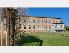 Großes, rechteckiges Gebäude mit beiger Fassade, zahlreichen rechteckigen Fenstern und einer Terrasse mit Bogenfenstern auf der rechten Seite; davor eine gepflegte Rasenfläche und links ein Gewächshaus mit rankenden Pflanzen. Der Himmel ist klar und blau.