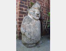 Verwitterte Steinfigur in Zwergform mit spitzer Mütze vor einer roten Backsteinmauer. Die Figur steht auf einem Betonboden, daneben sind einige Pflanzen und ein schmaler Holzpfahl zu sehen.