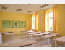 Ein gelber Raum mit mehreren Behandlungsliegen, die in zwei Reihen angeordnet sind. Große Fenster mit gelben Vorhängen und Wanddekoration in Form von grünen Bildern mit Kreismustern definieren den Raum.