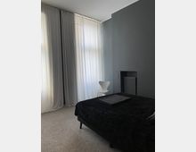 Schlafzimmer mit grauen Wänden und einem Teppichboden, ein Bett mit schwarzer Tagesdecke im Vordergrund, weiße Vorhänge vor hohen Fenstern und ein weißer Stuhl in der Ecke neben einem Heizkörper.