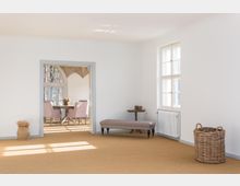 Heller Innenraum mit minimalistischem Design, bestehend aus einem leeren Vorraum mit einem Fenster, einer gepolsterten Sitzbank, einem kleinen Beistelltisch und einem Korb. Im Hintergrund ist ein Essbereich mit einem runden Holztisch, pastellfarbenen Stühlen und einem gotischen Bogenfenster sichtbar, durch das Licht einfällt.