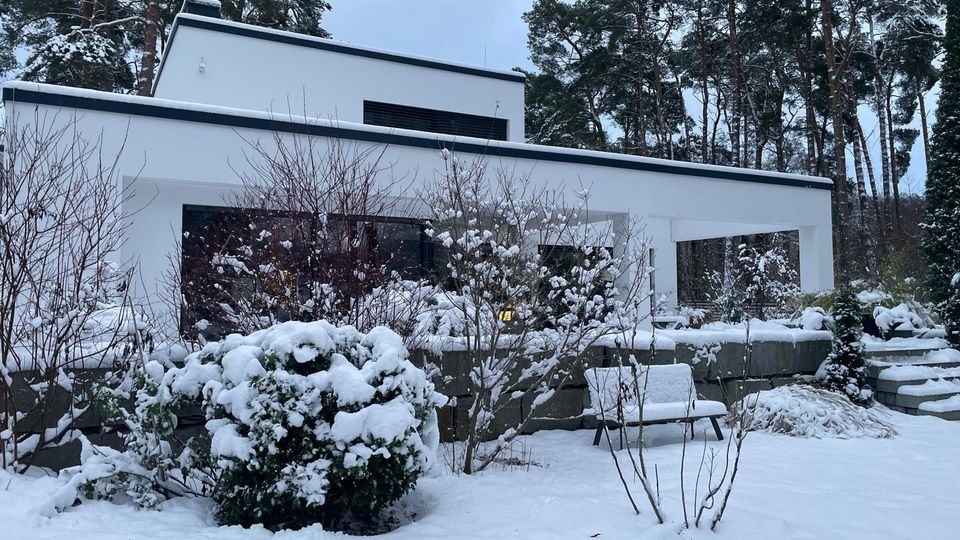 Modernes, weißes Flachdachhaus mit klaren, rechteckigen Formen und großer überdachter Terrasse, umgeben von hohen Kiefern. Im Vordergrund liegt ein verschneiter Garten mit Sträuchern, niedriger Steinmauer und einer Bank im Schnee.