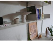 Weiße Wand mit einem Metallregal, das mehrere Regalbretter enthält. Auf den Brettern stehen Bücher, Keramikobjekte, eine Lampe, ein gerahmtes Bild mit geometrischen Mustern und kleine Dekorationsgegenstände.