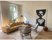 Helles Wohnzimmer mit einem großen gelben Ecksofa, zwei hellen Sesseln und einem runden Couchtisch. An der Wand hängt ein monochromes Gemälde einer Frau, daneben ein großes Fenster mit Blick ins Grüne; der Boden besteht aus Fischgrätparkett.