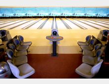 Bowlingbahn mit mehreren Bahnen, umgeben von blau gepolsterten, grauen Sitzschalen auf beiden Seiten. Im Vordergrund befindet sich eine Bedienkonsole und ein Bereich mit Teppichboden.