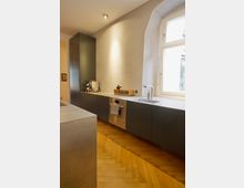 Moderne Küchenzeile mit dunklen Schränken und integrierten Geräten, darunter ein Backofen. Eine Spüle befindet sich unter einem großen, bogenförmigen Fenster. Der Boden besteht aus Holz in Fischgrätmuster.