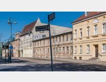 Straßenszene mit teils renovierten, teils verfallenen Gebäuden, darunter ein gelbes Wohnhaus und ein älteres Gebäude mit abblätternder Fassade und Graffiti. Im Vordergrund steht ein Straßenschild mit den Namen „Goepelstraße“ und „Berliner Straße“, sowie Straßenlaternen und Schienen für den Straßenbahnverkehr.