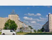 Straßenszene mit einem öffentlichen Platz inmitten historischer Gebäude im sozialistischen Baustil, flankiert von symmetrischen Wohn- und Geschäftsgebäuden. In der Mitte befindet sich ein großer Springbrunnen, umgeben von Grünflächen, vor einem klaren Himmel mit Wolken.