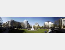 Eine städtische Parkanlage mit einer zentralen Springbrunnenanlage, umgeben von modernen und historischen Gebäuden. Im Vordergrund erstreckt sich eine gepflegte Rasenfläche, gesäumt von Blumenbeeten, Bänken und Bäumen.