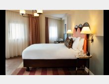 Ein stilvolles Schlafzimmer mit einem großen Bett und gepolstertem, rosa Kopfteil, flankiert von zwei Nachttischen mit Lampen. Im Hintergrund befinden sich Vorhänge, große Fenster mit transparenten Gardinen und ein kleiner Tisch mit Stuhl.