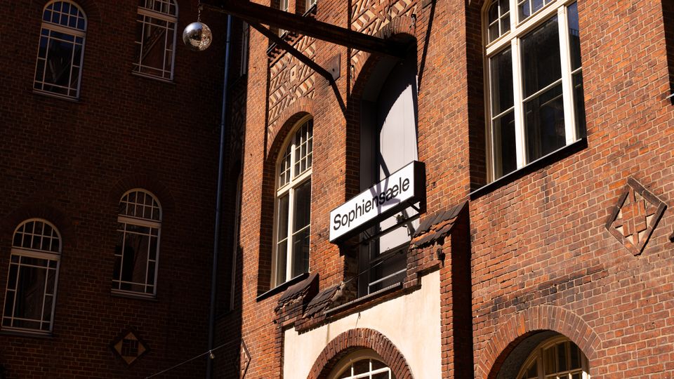 Backsteinfassade eines mehrstöckigen Gebäudes mit hohen, bogenförmigen Sprossenfenstern und dekorativen Mauerwerksmustern. An einem Metallausleger hängt ein Schild mit der Aufschrift „Sophiensæle“ über einem Eingang.
