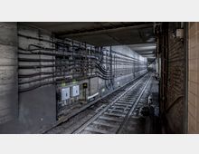 Unterirdischer Eisenbahntunnel mit Schienen, Kiesbett und Kabeln, die an den Betonwänden befestigt sind. Die Szenerie zeigt eine Mischung aus technischer Infrastruktur und engen Tunnelräumen mit Beleuchtung in der Ferne.