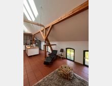 Wohnraum in einem Dachgeschoss mit Holzbalken, roten Fliesenboden und schräger Decke. Zu sehen sind ein Küchenbereich mit weißer Kochinsel, eine schwarze Liege, ein Beistelltisch mit Lampe, zwei kleine Fenster mit Blick ins Grüne und ein gemusterter Hocker auf einem flauschigen Teppich.