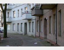 Straßenansicht eines Wohngebäudes mit hellblauer und beige-brauner Fassade, mehreren Balkonen und rechteckigen Fenstern. Der Gehweg ist gepflastert, teils mit Laub bedeckt, und ein Baum steht nahe an der Häuserwand.