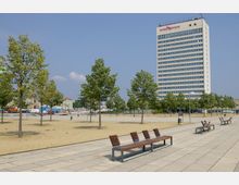 Weitläufiger öffentlicher Platz mit gepflasterten Wegen, einzelnen Reihen von Bänken und jungen Bäumen. Im Hintergrund steht ein modernes, mehrstöckiges Hotelgebäude mit der Aufschrift „hotel Mercure“.