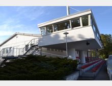 Gebäude eines Rad- und Reitstadions mit weißer Fassade, großen Fenstern im oberen Bereich und einer Außentreppe aus Metall. Unterhalb befinden sich überdachte Sitzreihen mit roten Kunststoffstühlen, daneben wächst Grünfläche.