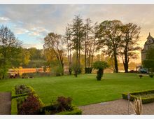 Grünanlage mit gepflegtem Rasen und blühenden Beeten im Vordergrund, umgeben von Bäumen am Ufer eines ruhig daliegenden Sees bei Sonnenuntergang. Rechts im Bild ist ein historisches Gebäude mit einer Verzierung am Dach zu sehen.