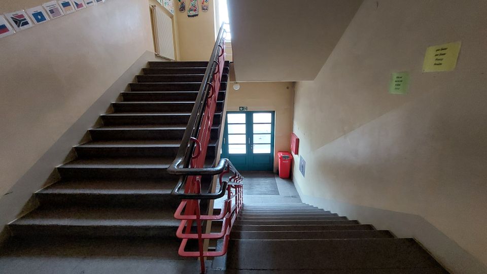 Treppenhaus in einem Innenbereich mit beigen Wänden, dunkelbraunen Stufen und einem roten Geländer. Am unteren Ende ist eine Glastür zu sehen, daneben ein roter Abfalleimer und ein Feuerlöscher. An den Wänden hängen Hinweiszettel und einige Dekorationen.