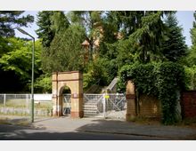 Gelbes Backsteinportal mit rundem Bogen und Metallgittertor, flankiert von Mauerteilen mit Kletterpflanzen, eingerahmt von dichter Baumvegetation; im Hintergrund ein historisches Gebäude mit roten Dächern. Eine Straße und eine Straßenlaterne befinden sich im Vordergrund.