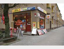 Eckladen in einem mehrstöckigen Wohnhaus in einer Stadt, mit einer Backwaren- und Zeitschriftenauslage in den Schaufenstern. Vor dem Eingang stehen Werbetafeln und Schilder, daneben Gehweg und kahle Bäume.