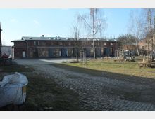 Ein gepflasterter Hof mit einem flachen, langgestreckten Ziegelgebäude im Hintergrund, das zahlreiche Fenster und Türen besitzt. Im Vordergrund sind Bäume, kahle Sträucher und ein Bereich mit Gras zu sehen.