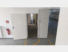 Innenansicht eines einfachen, weiß gestrichenen Gebäudeflurs mit geöffneten Türen, die in einen weiteren Raum führen. Links ist ein Feuerlöscher an der Wand angebracht, daneben ein Heizkörper, der Boden besteht aus Beton mit gelben Markierungen.