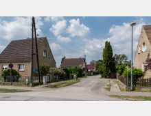 Straßenszene in einem Wohnviertel mit Einfamilienhäusern, die verputzte Fassaden und Satteldächer haben. Links und rechts der Straße befinden sich Vorgärten mit Zäunen, hohen Bäumen und Sträuchern, bei teilweisem bewölkten Himmel.