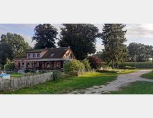 Backsteinbauernhaus mit Satteldach und grünen Fensterrahmen in ländlicher Umgebung, umgeben von Bäumen, Sträuchern und einem Holzzaun; im Vordergrund eine Wiese und ein unbefestigter Weg.