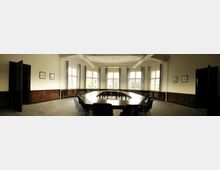 Ein rechteckiger Konferenzraum mit großen Fenstern, die Tageslicht einlassen. In der Mitte befindet sich ein großer, u-förmiger Holztisch, umgeben von schwarzen Stühlen; die Wände sind holzvertäfelt und mit gerahmten Bildern dekoriert.