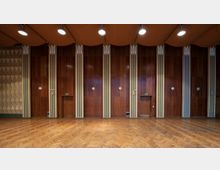 Großer Saal mit Holzwänden in warmem Braunton und vertikalen Streifen als Dekorelemente. Der Boden ist mit Fischgrätparkett ausgelegt, die Decke ist mit einer orangefarbenen Verkleidung sowie hängenden Lampen gestaltet.