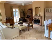 Wohnzimmer mit klassisch-eleganter Einrichtung, beigefarbenen Wänden und einem Fischgrät-Parkettboden. Der Raum enthält ein cremefarbenes Sofa, Holzmöbel, einen Marmorkamin mit Spiegel, sowie doppelflügelige Glastüren, die auf eine Terrasse führen.