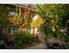 Ein zweistöckiges Wohnhaus mit Ziegeldach, dessen Fassade dicht mit Efeu und herbstlich gefärbten Kletterpflanzen bewachsen ist. Davor liegt ein begrünter Innenhof mit Bäumen, Pflanzenkübeln, Treppenstufen und einer kleinen Sitzgruppe aus Holztisch und Stühlen.